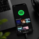 A mobile phone currently on the spotify app homepage beside a laptop